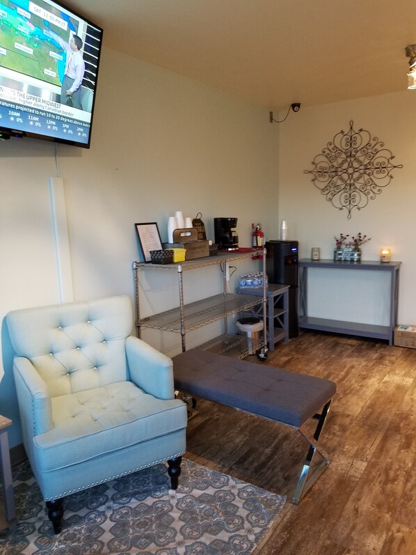 A living room with a light blue armchair, a grey bench, a coffee station, a TV mounted on the wall, and a decorative wall piece. - SureStay by Best Western Yerington (Yerington)