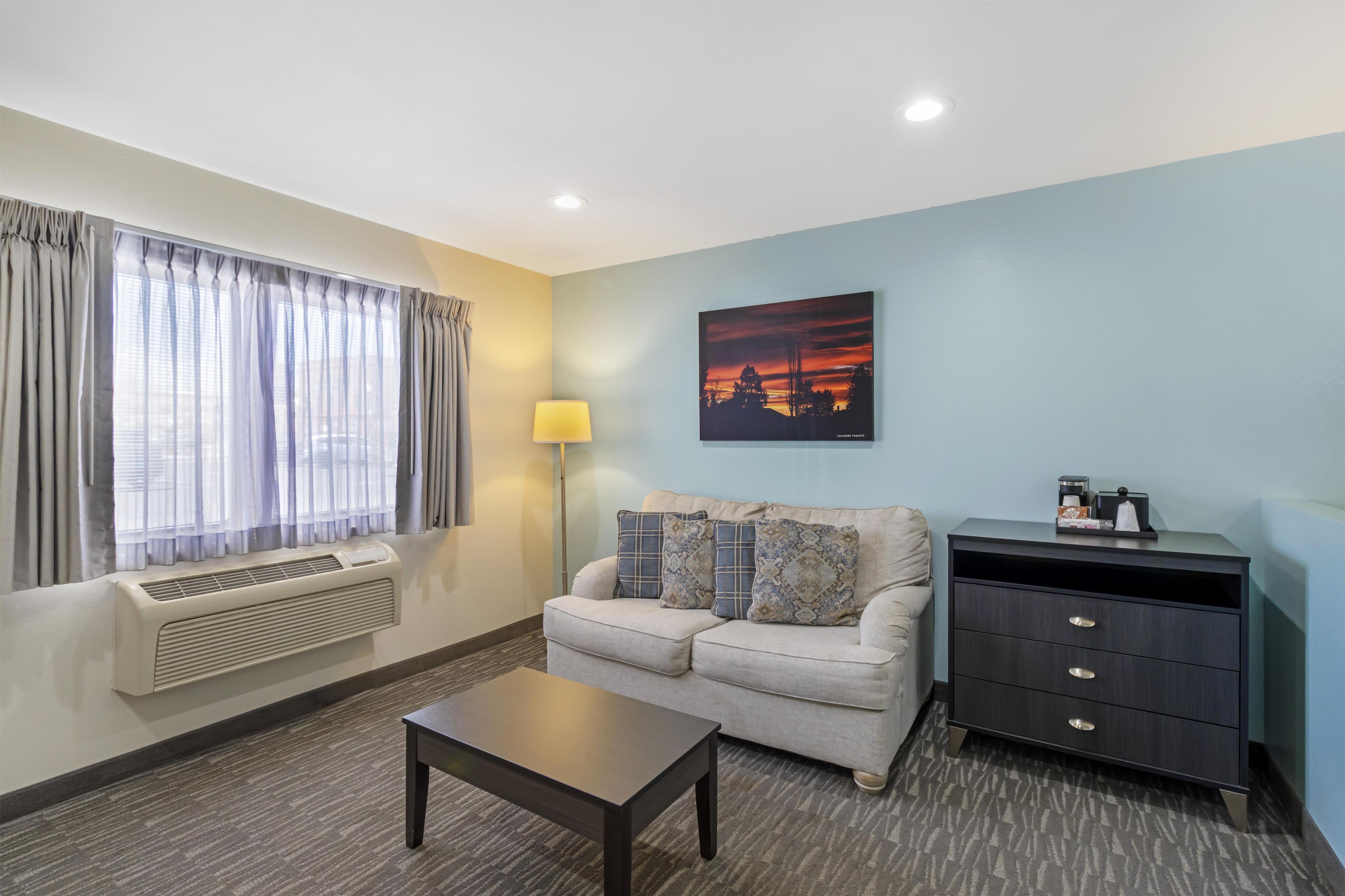 A hotel room with a sofa, a table, a lamp, a painting, and a desk with drawers.