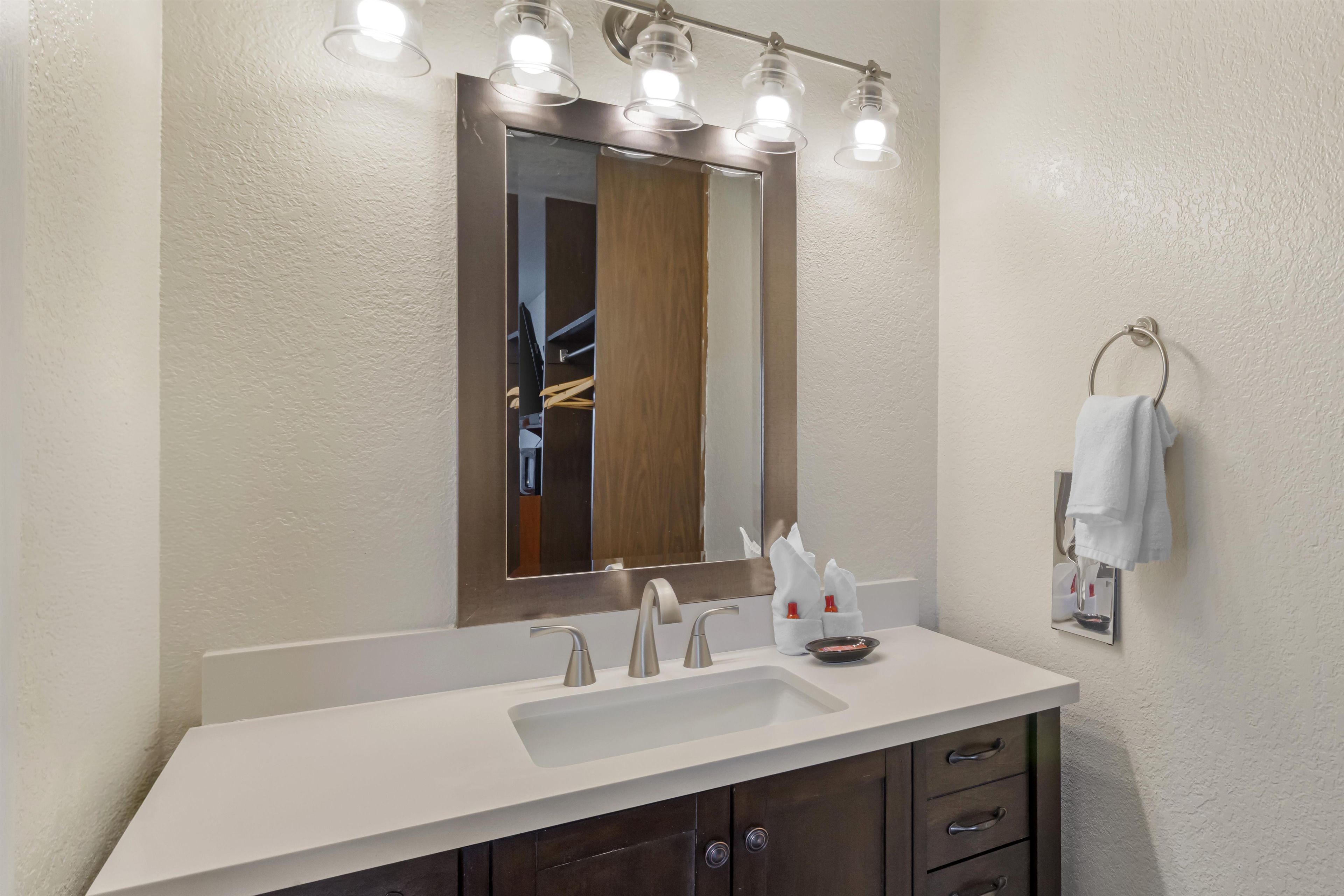 Une salle de bain avec un lavabo blanc, deux robinets, un miroir avec éclairage intégré, un porte-serviettes et un meuble avec des tiroirs.