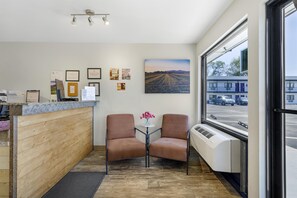 A reception area with a wooden counter, two leather chairs, a small table with a vase of flowers, and a large window with a view of a parking lot and buildings. - SureStay by Best Western Yerington (Yerington)