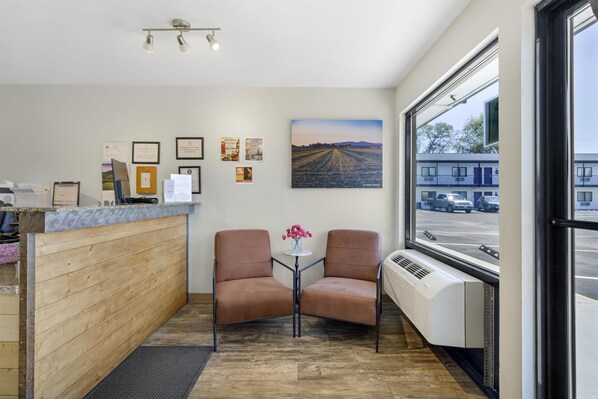 Una zona de recepción con mostrador de madera, dos sillones de cuero, una mesa pequeña con un florero y un ventanal con vistas a un estacionamiento y edificios.