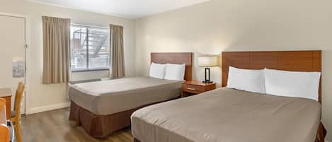 A hotel room with two beds, a desk, a chair, a window with curtains, and a nightstand with a lamp.