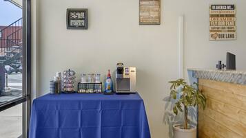 Une pièce avec une table recouverte d’une nappe bleue, une machine à café et divers récipients. On peut voir des panneaux d’encouragement au mur et une plante en pot.