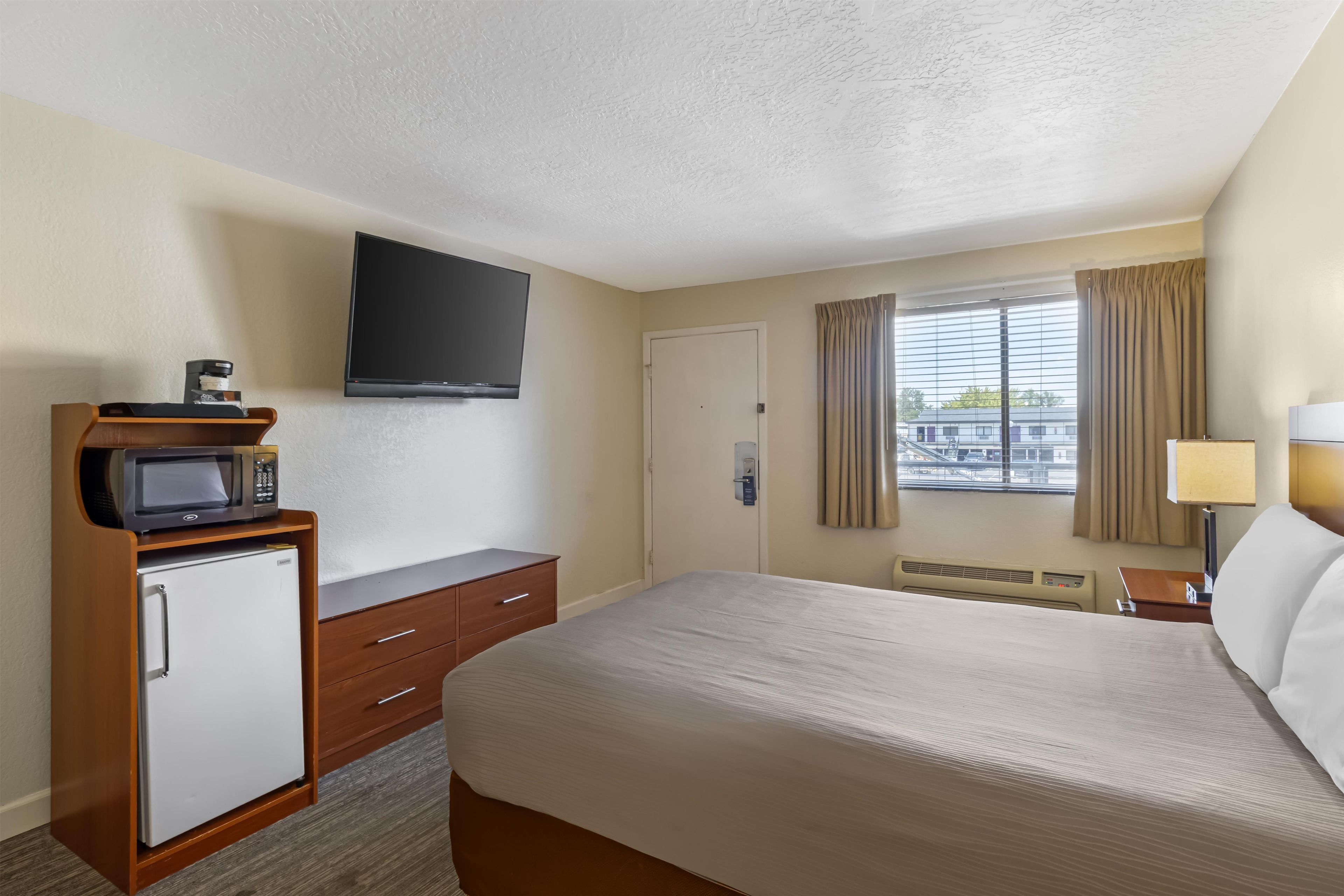 A hotel room with a bed, a television mounted on the wall, a minibar, a desk, and a window with curtains.