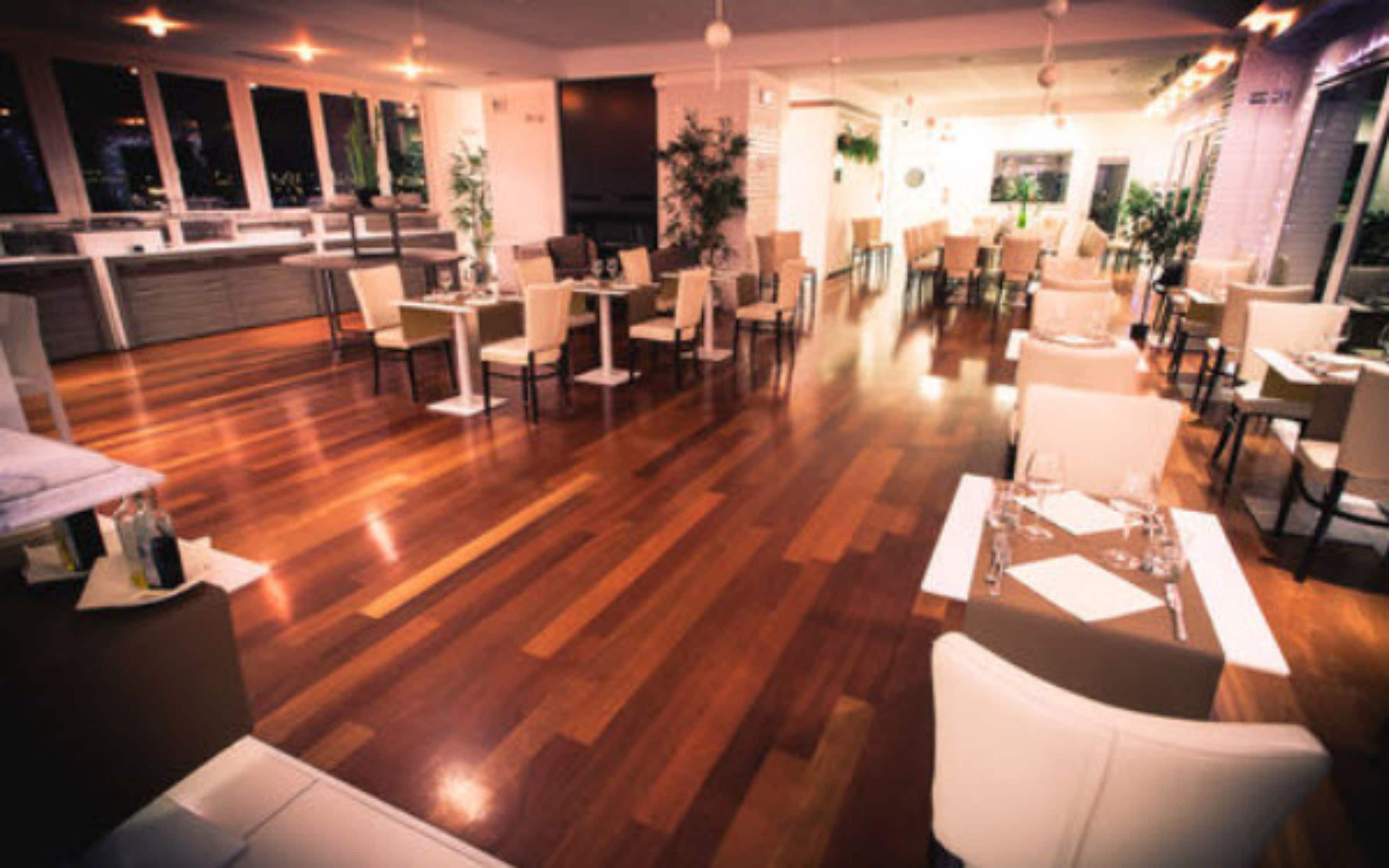Um restaurante espaçoso com piso de madeira, mobiliário branco e amplas janelas.