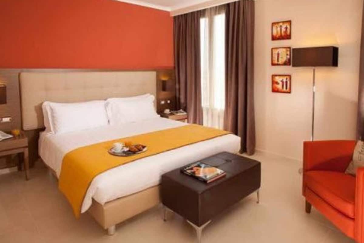 A hotel room with a large bed, a bedside table, a red armchair, and a floor lamp.