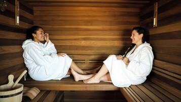 Two individuals in a sauna, seated on wooden benches with towels wrapped around their legs.