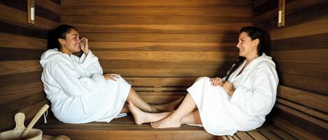 Two individuals in a sauna, seated on wooden benches with towels wrapped around their legs.