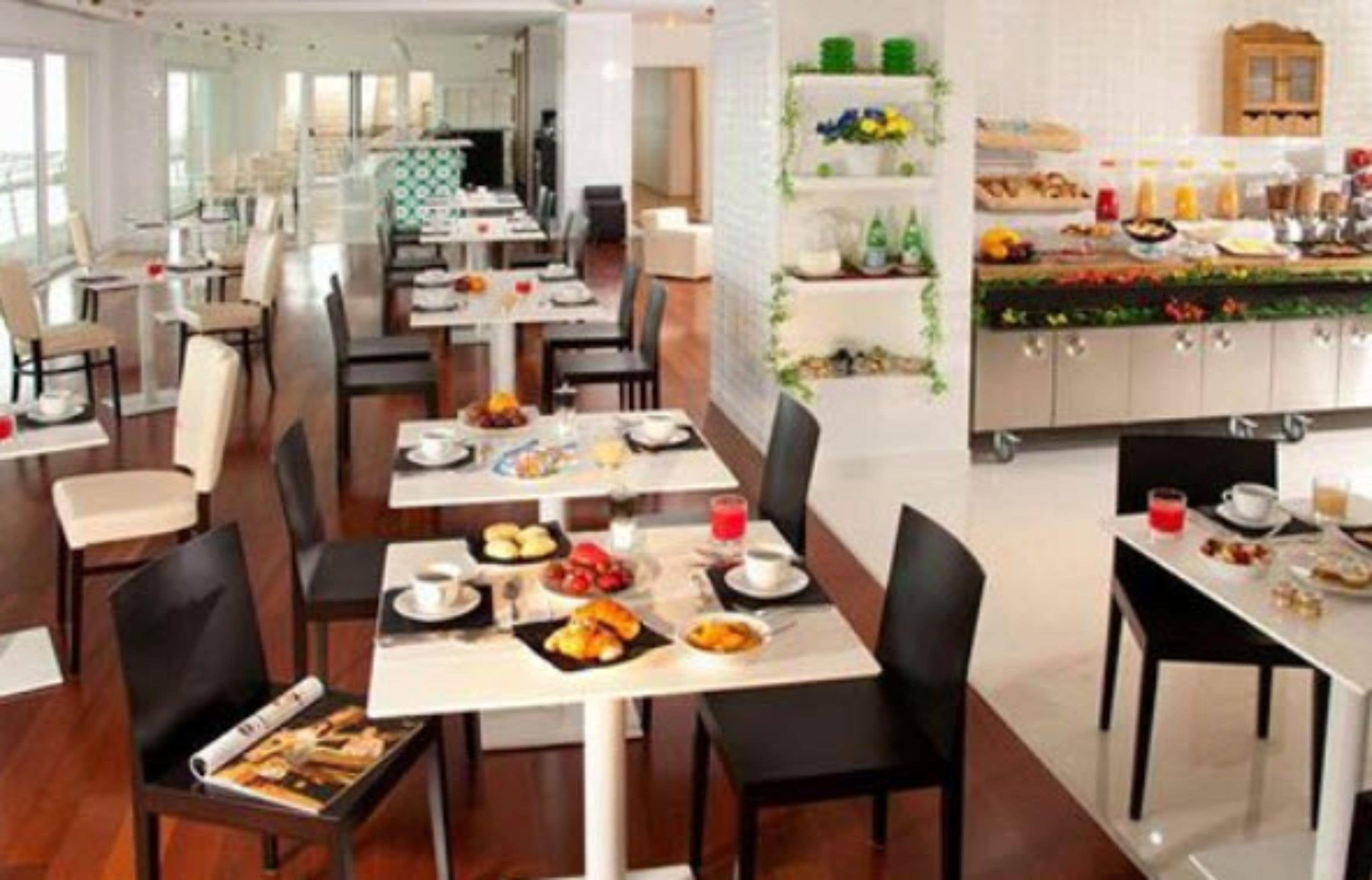 Zona ristorazione di un hotel con diversi tavoli e sedie, angolo cottura con buffet e vista sulla sala da pranzo attraverso ampie finestre.