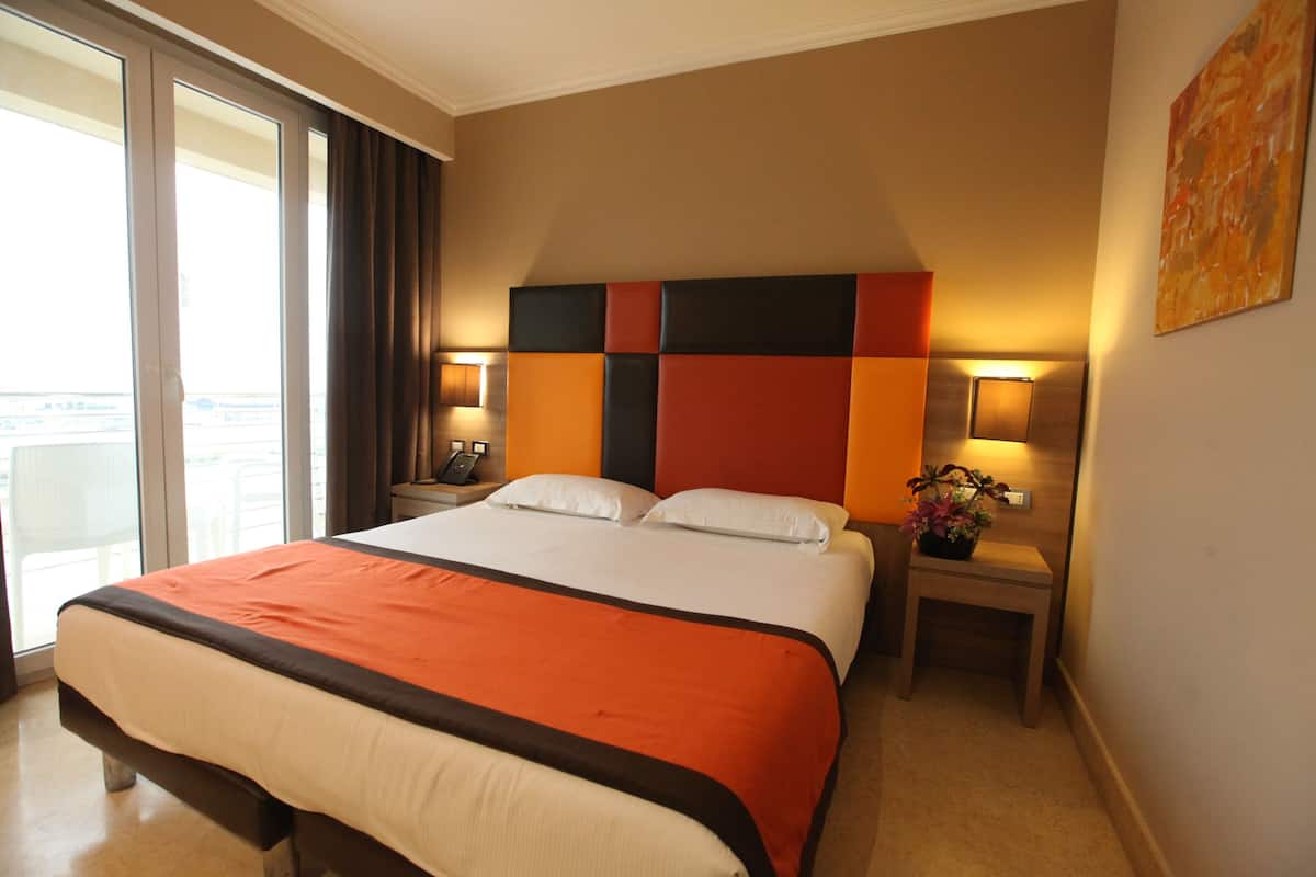 A hotel room with a large bed, a bedside table with a lamp, a nightstand with a phone, and a window with curtains.