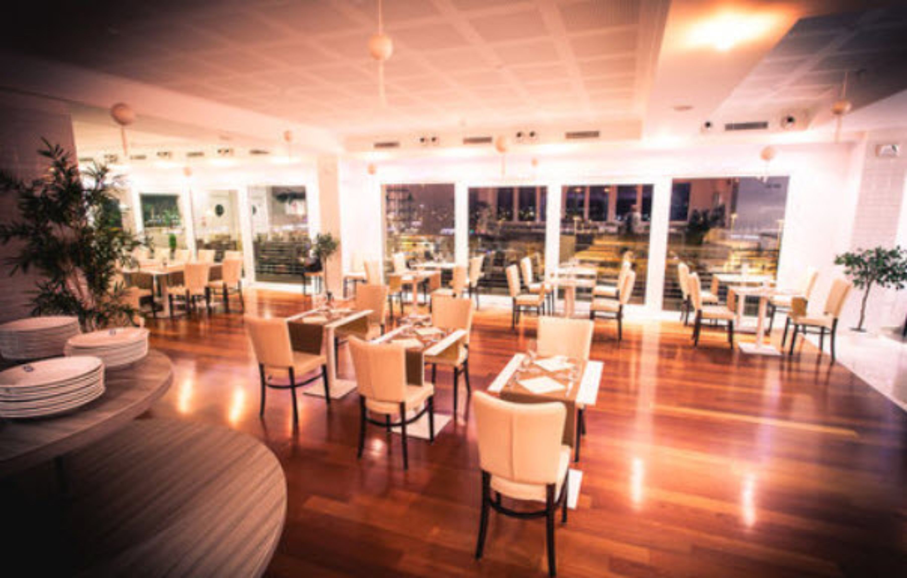 A spacious restaurant with multiple tables and chairs, large windows offering a city view, and a polished wooden floor.