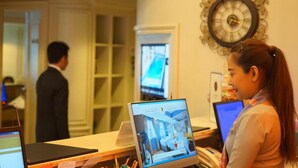 Un mostrador de recepción con una computadora portátil mostrando una vista de la habitación, un reloj en la pared y un hombre al fondo.