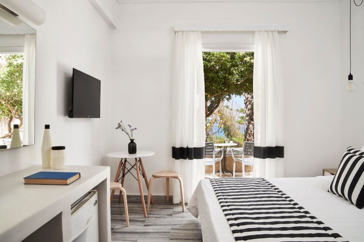 A hotel room with a bed, a desk, a chair, a small table with a vase, and a TV.
