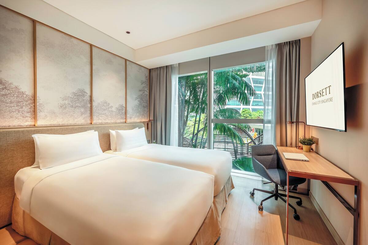 Ein Hotelzimmer mit zwei Betten, einem Schreibtisch, einem Stuhl, einem Fernseher und einem großen Fenster mit Blick auf Grünflächen und ein Gebäude.