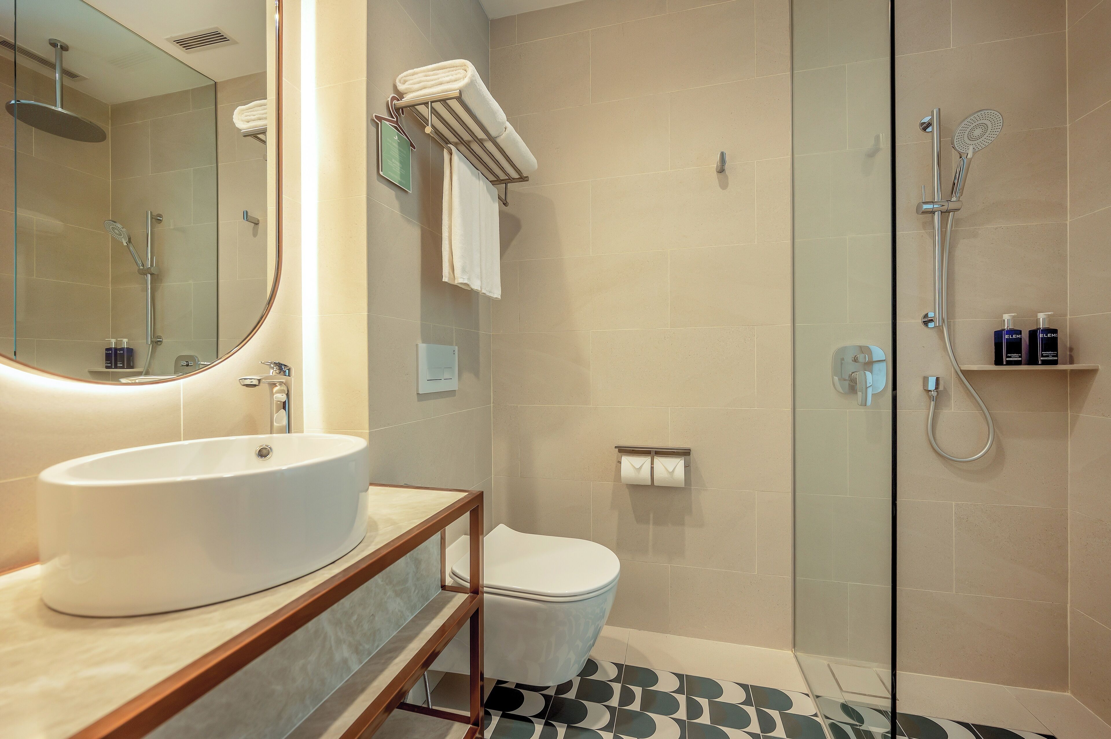 Une salle de bain moderne avec un grand miroir, un lavabo rond, des toilettes et une douche séparée par une paroi de verre.