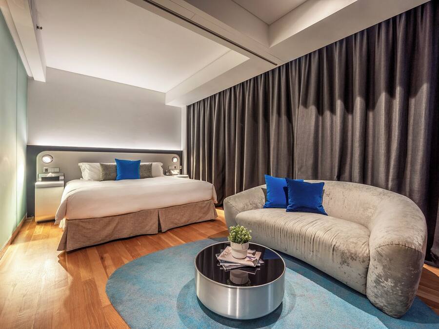 A modern hotel room with a large bed, a sofa, a coffee table, and a large window with curtains.