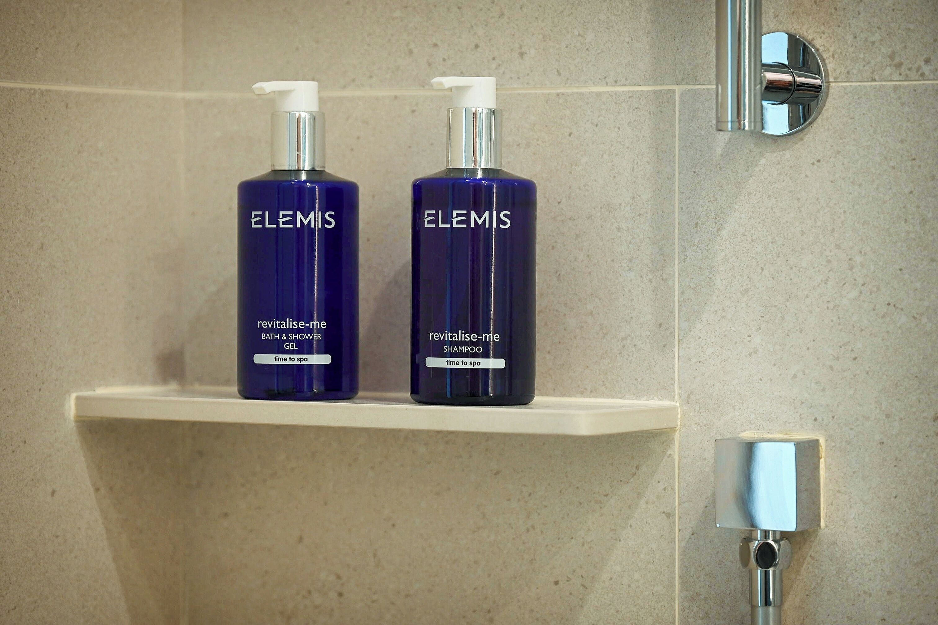 Two bottles of Elemis Revitalise-Me Bath & Shower Gel and Shampoo on a white shelf in a bathroom with a tiled wall and a chrome faucet.