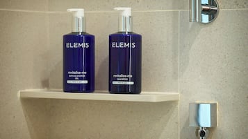 Two bottles of Elemis Revitalise-Me Bath & Shower Gel and Shampoo on a white shelf in a bathroom with a tiled wall and a chrome faucet.