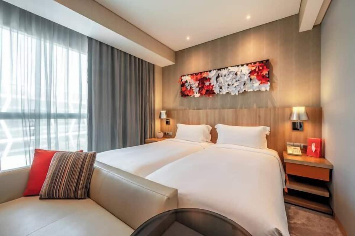 Hotellihuone, jossa on kaksi sänkyä, sohva, suuri ikkuna verhoilla, seinälle kiinnitetty taideteos, yöpöytävalaisimet ja pieni pöytä, jossa on puhelin.