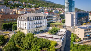 A cityscape with a mix of traditional and modern buildings, a prominent white building with a grey roof, and a modern glass building. - Hôtel Alpes & Lac (Neuchatel)