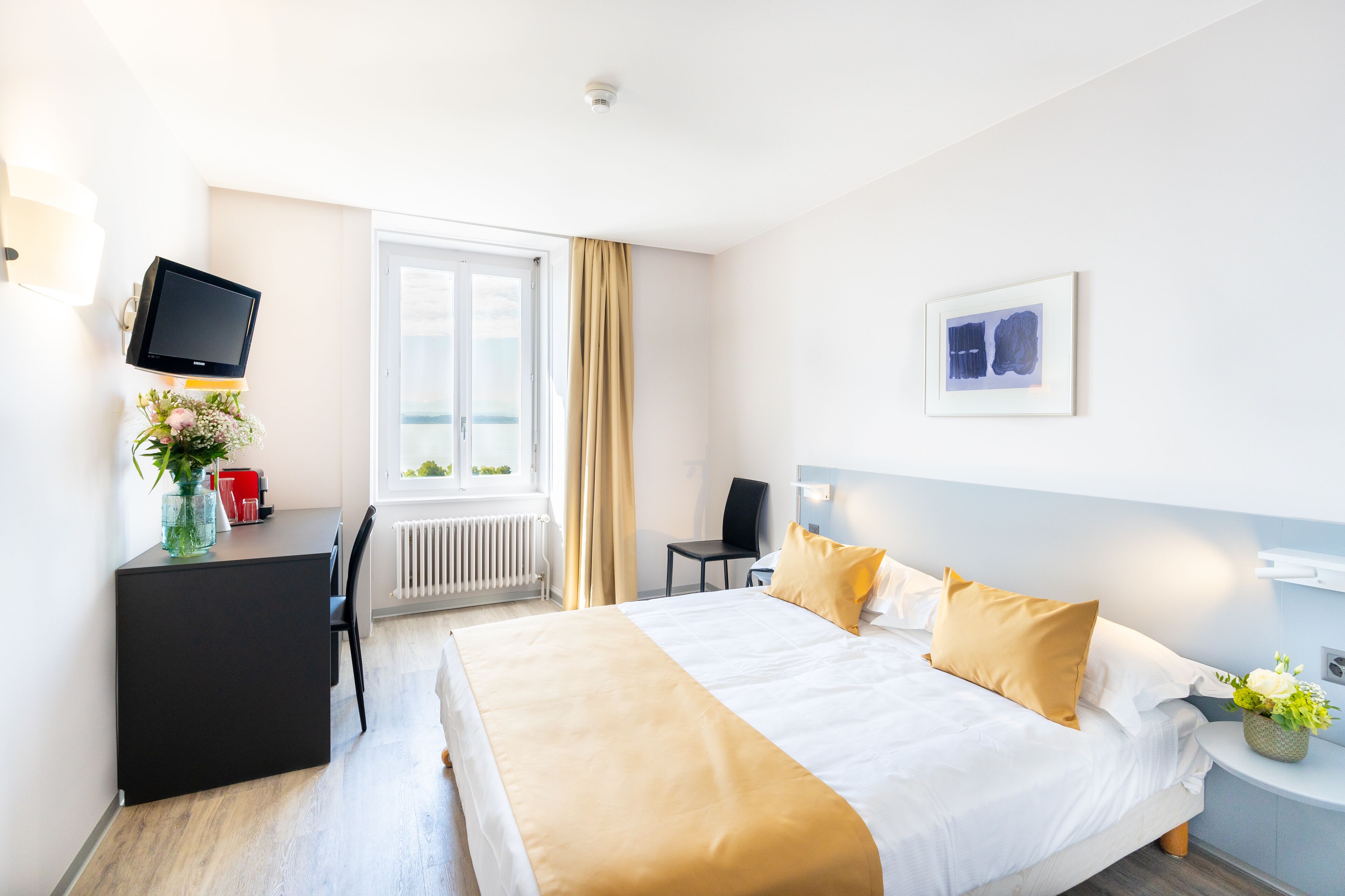 Ein Hotelzimmer mit einem Bett, einem Schreibtisch, einem Stuhl, einem Fernseher, einem Fenster mit Vorhängen und einem Wandbild.