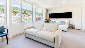A modern hotel room with a large window, a sofa, a bed, and a flat-screen TV. - Hôtel Alpes & Lac (Neuchatel)