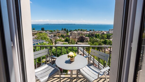 A balcony with a table and chairs, overlooking a cityscape and a body of water. - Hôtel Alpes & Lac (Neuchatel)