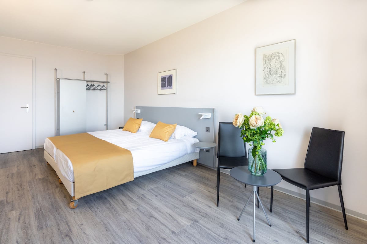 Una habitación de hotel con una cama, una silla, una mesita y un jarrón con flores.