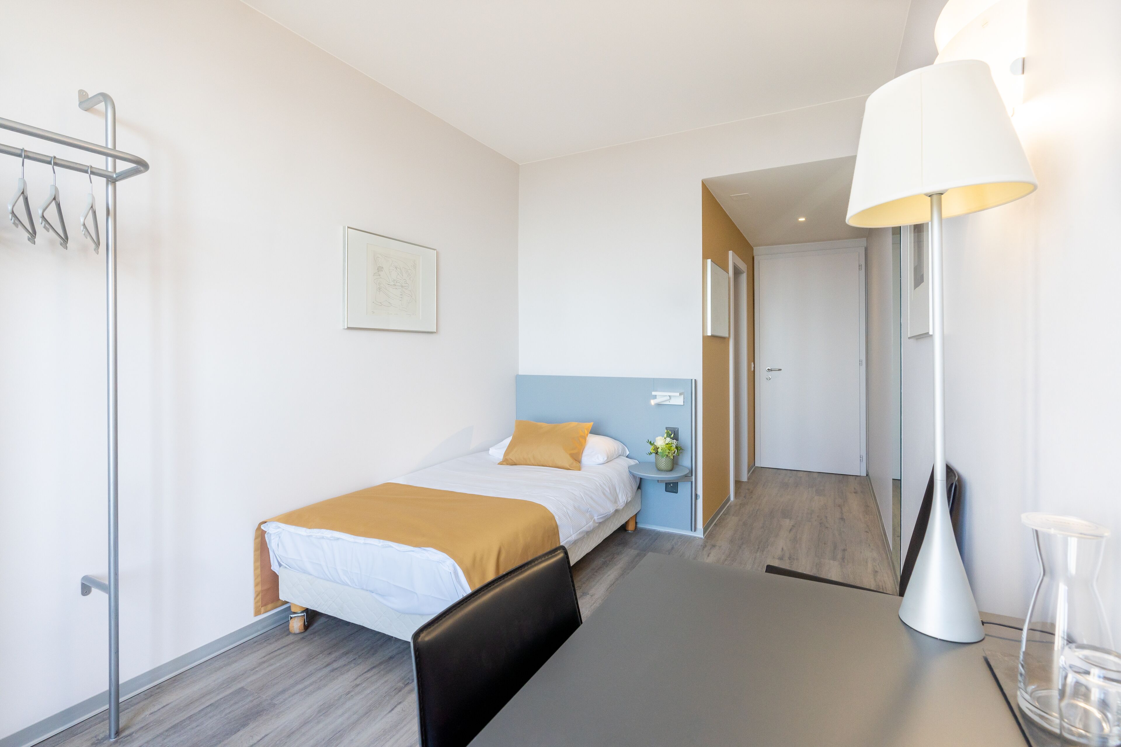 A modern hotel room with a bed, bedside table, lamp, and a view of the hallway.