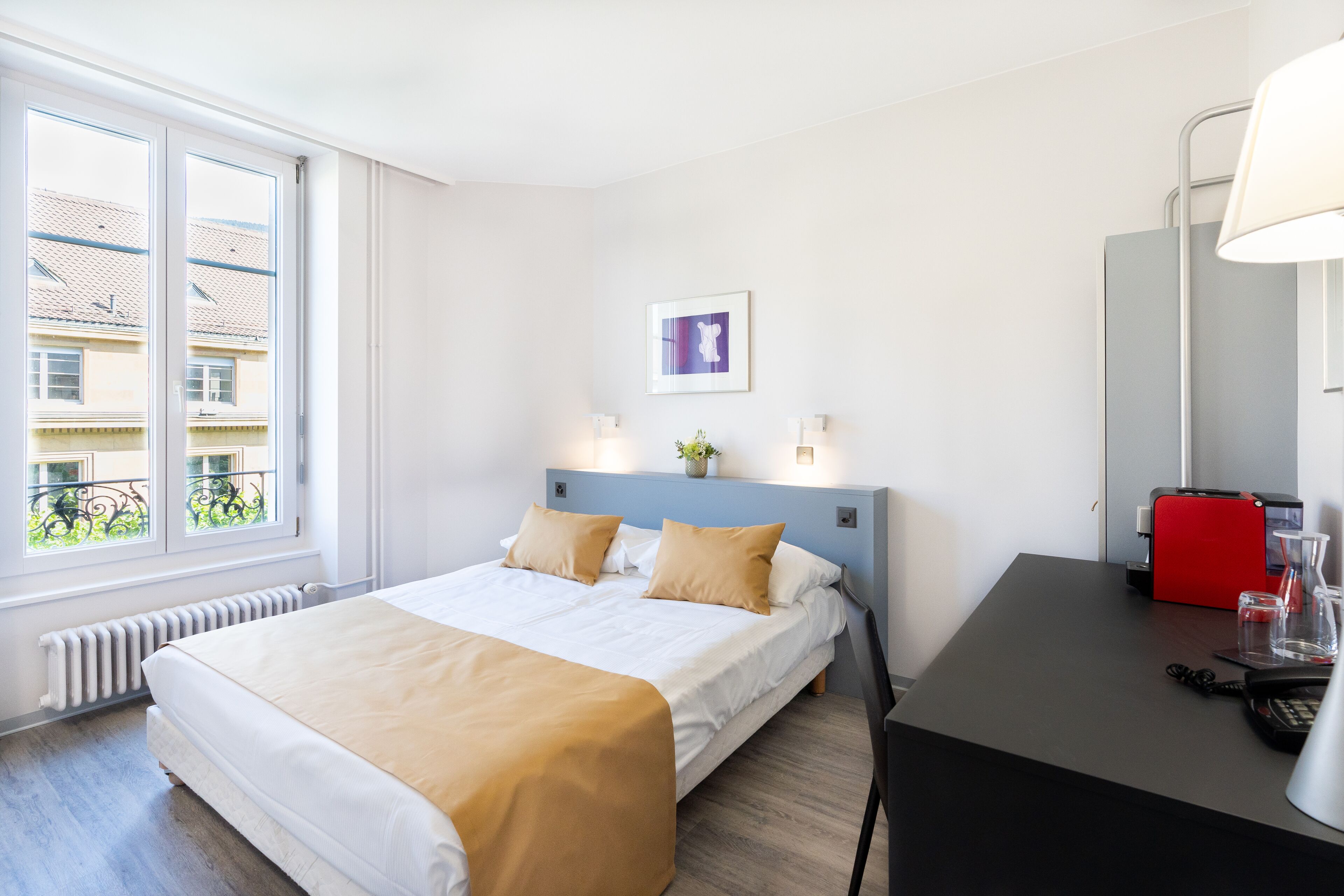 Ein Hotelzimmer mit einem Bett, einem Schreibtisch mit einem roten Telefon und einem Fenster mit Blick auf Gebäude.