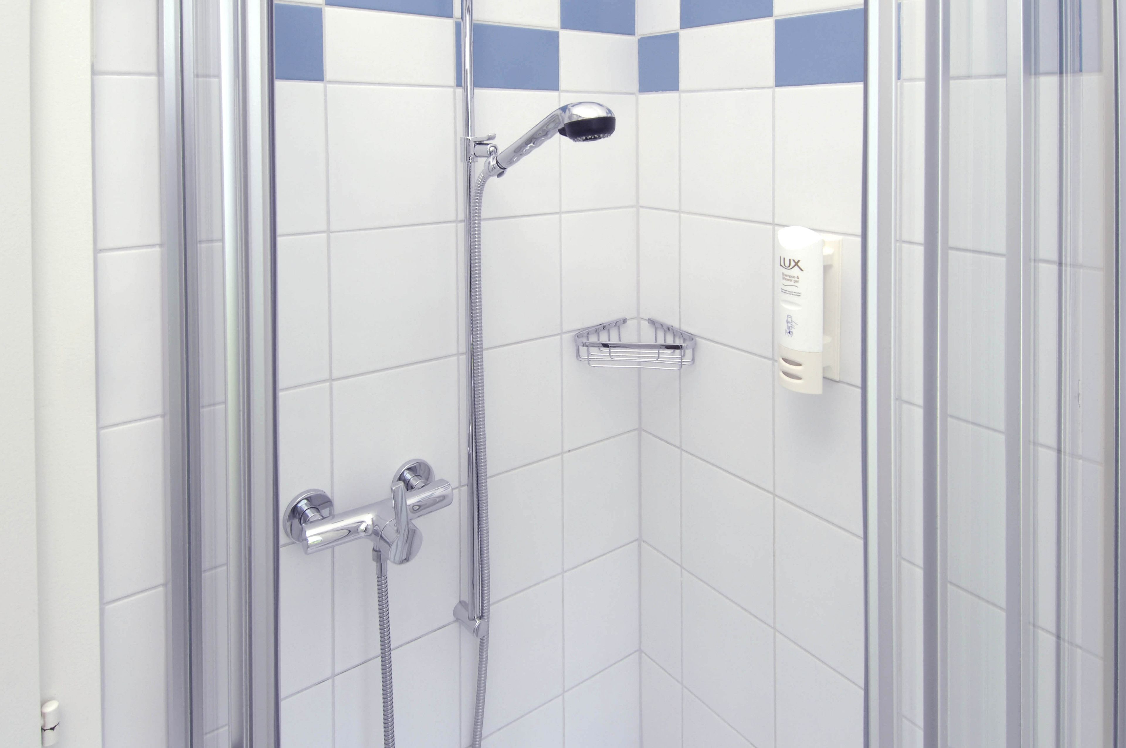 A shower with a handheld showerhead and a wall-mounted faucet, a soap dispenser, and a small shelf.
