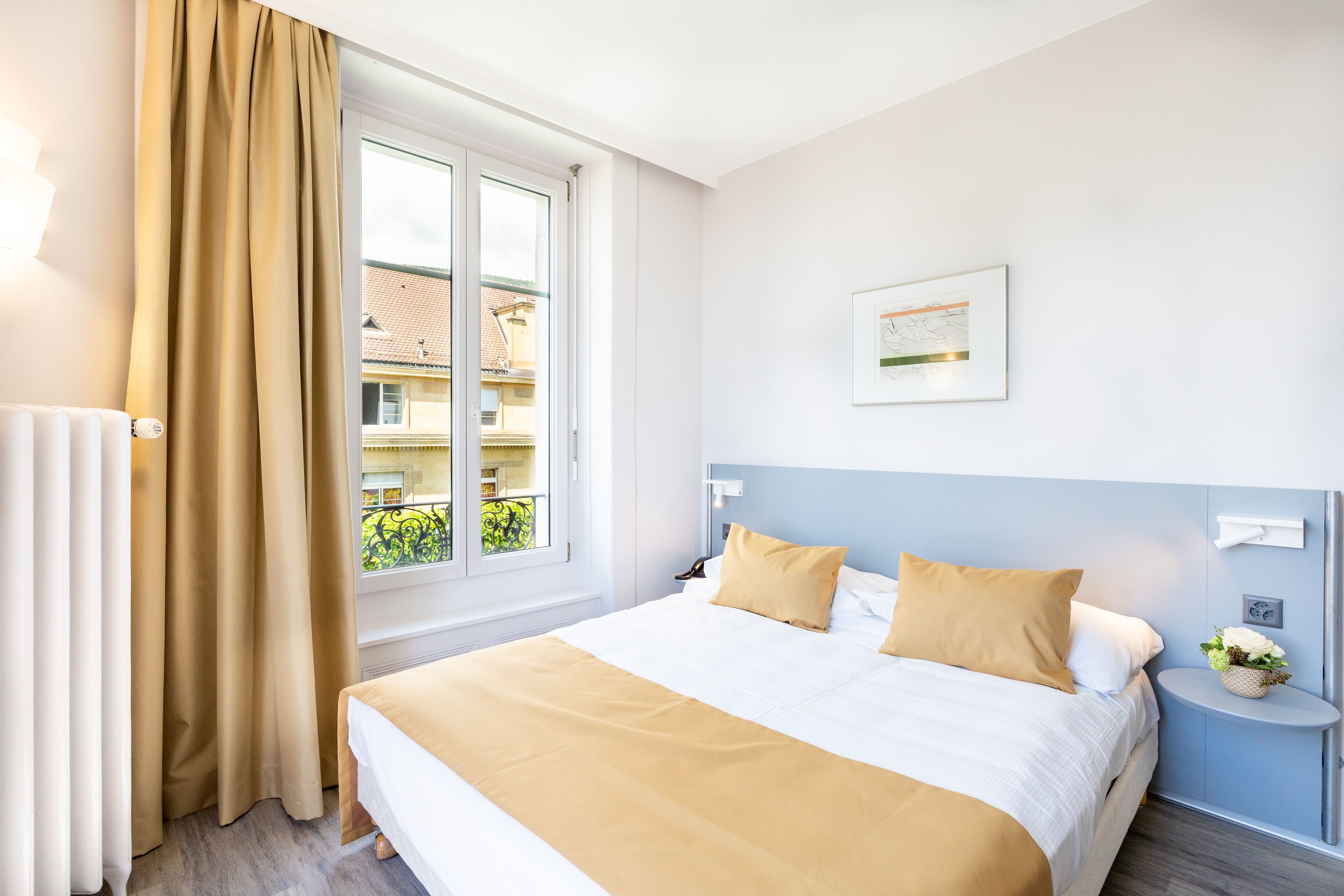 Una habitació d'hotel amb un llit gran, una finestra amb cortines, una tauleta de nit amb una làmpada i un quadre penjat a la paret.