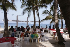 An outdoor dining area with white plastic chairs and tables, palm trees, and a view of the sea. - La Voile d'Or (Dakar)