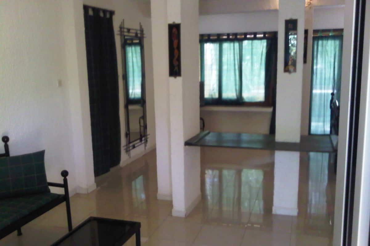 A spacious living area with a sofa, a coffee table, and a dining table. There are windows with curtains, a tiled floor, and a door leading to another room.