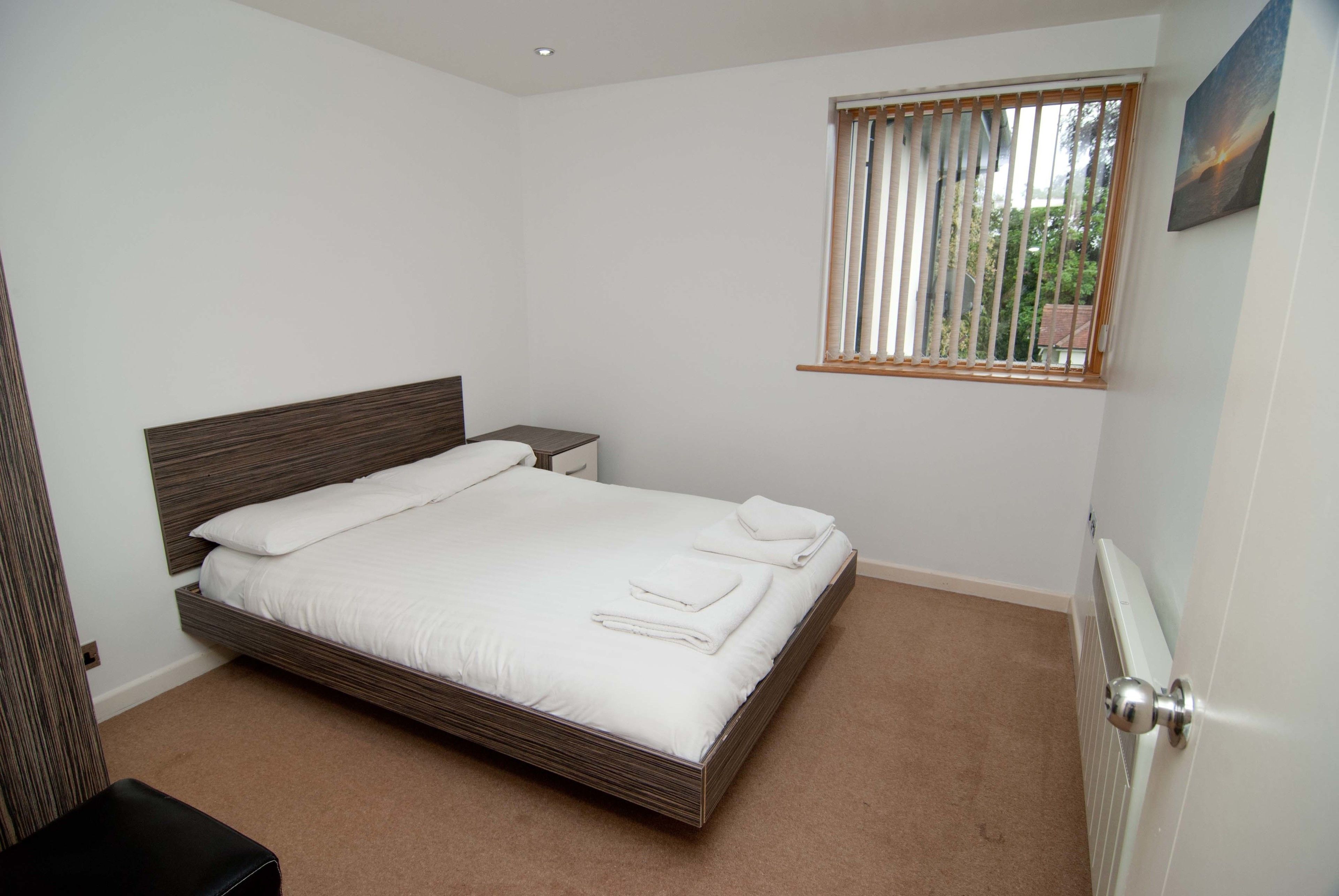 A bedroom with a single bed, a bedside table, a window with blinds, and a radiator.