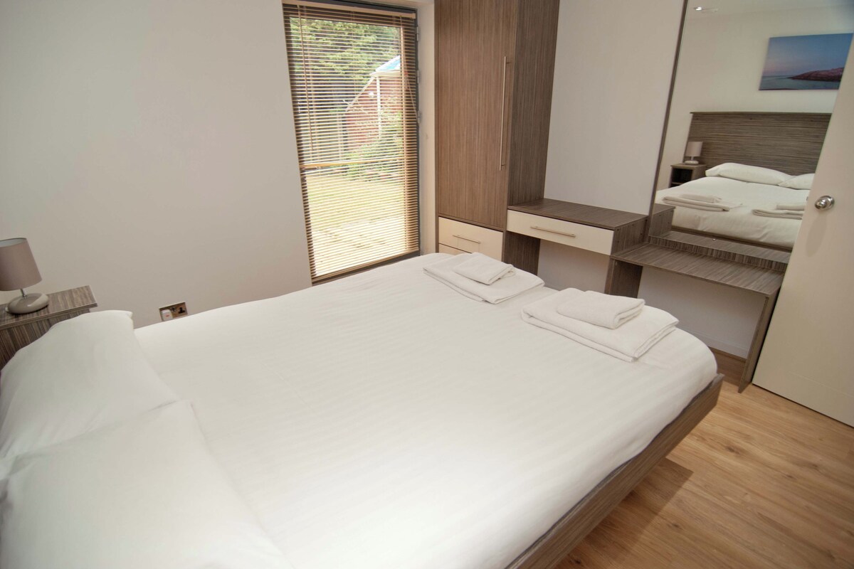A neatly made bed with white linens and towels, a wooden wardrobe, a small bedside table with a lamp, and a window with blinds.