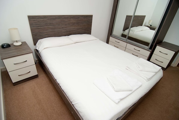 A neatly made bed with white linens and towels, a bedside table with a lamp and phone, and a large mirror reflecting the bed and room.