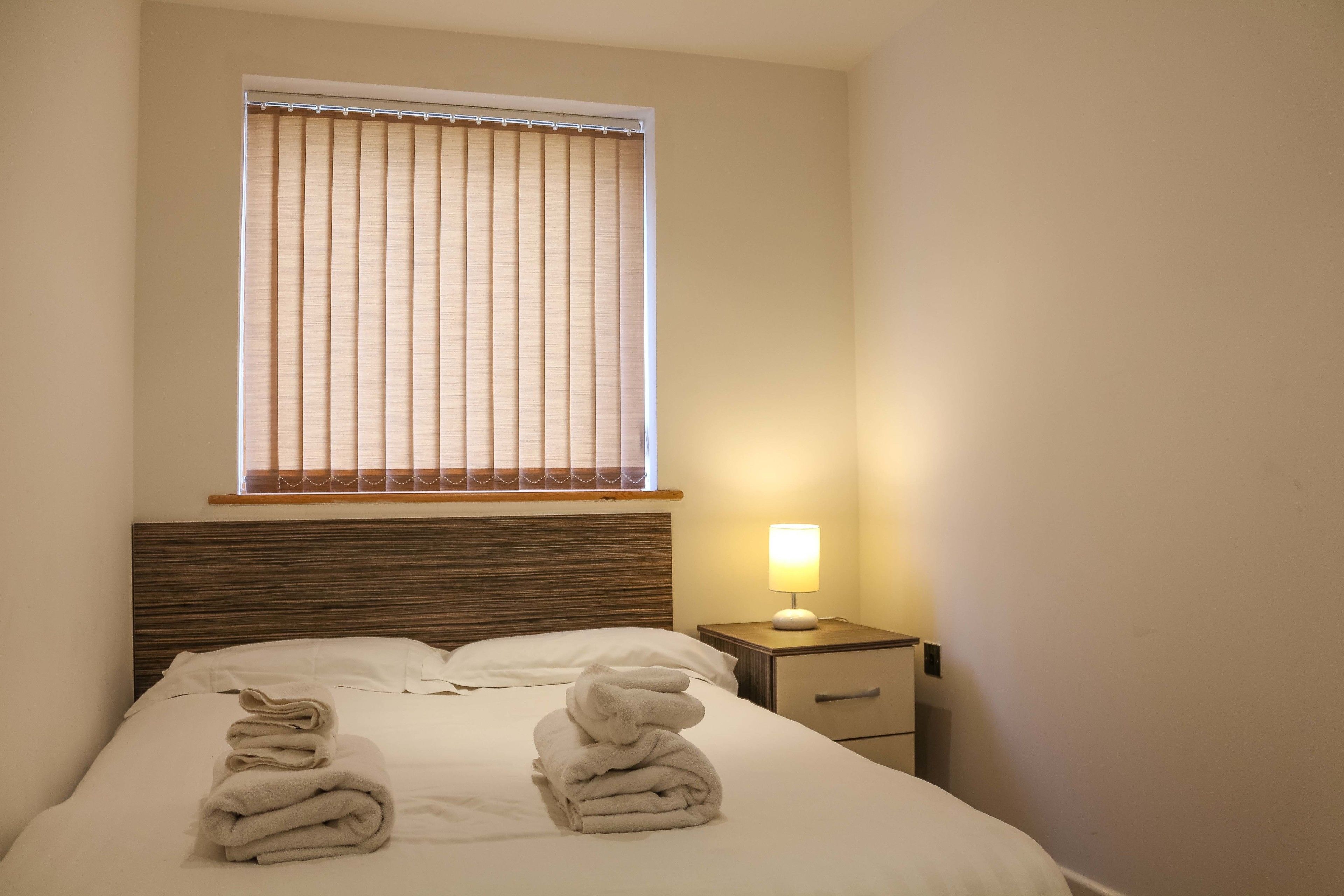 A neatly made bed with folded towels, a bedside lamp, and a window with blinds.