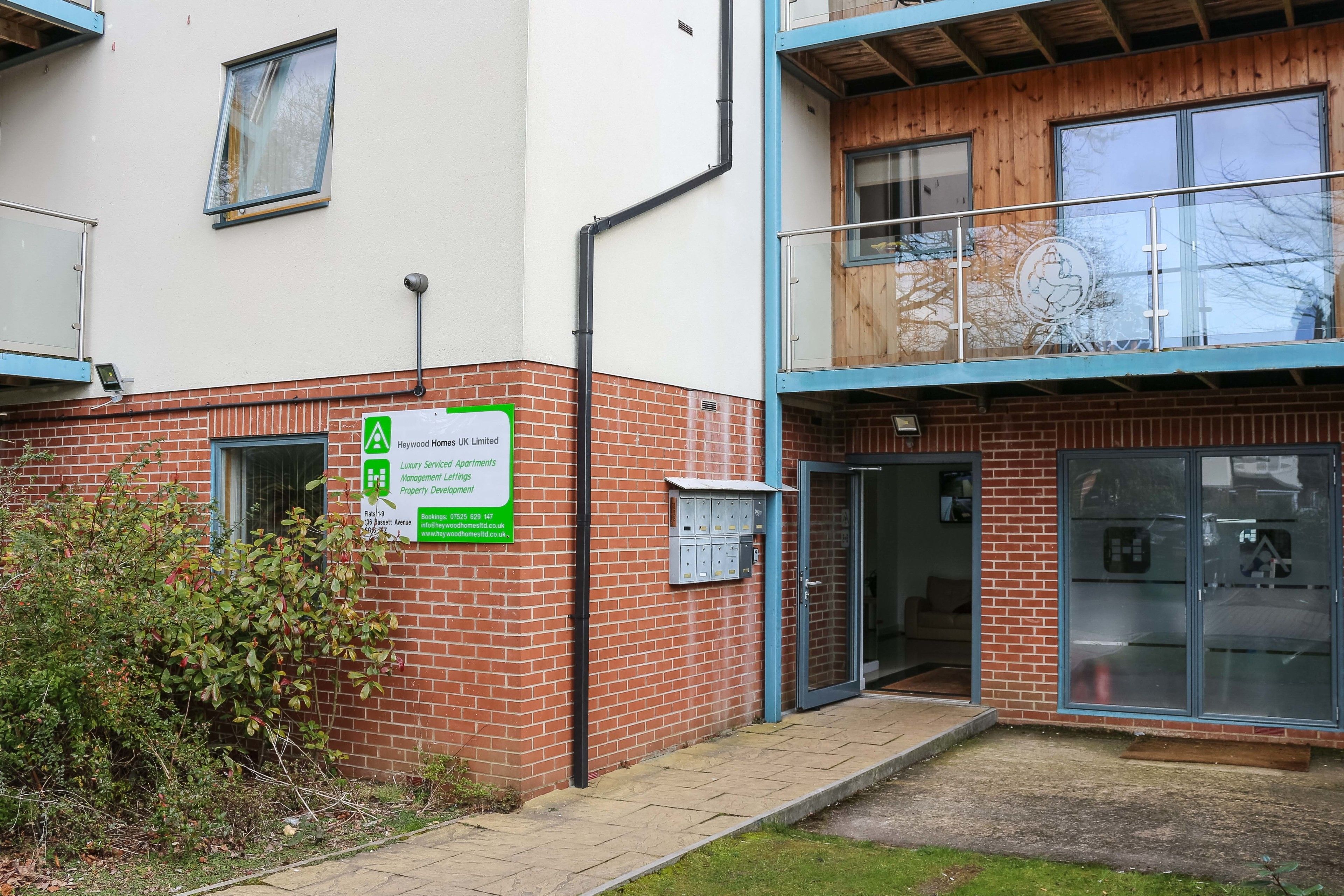 Un edificio con un cartel de Heywood Homes UK Limited, que indica apartamentos de lujo. Se observa una pared de ladrillo, un pequeño jardín y una entrada con puerta de cristal.