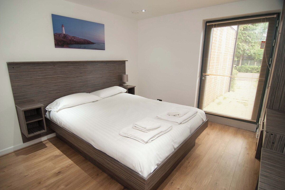 A bedroom with a large bed, a nightstand, a wall-mounted lamp, and a window with blinds.