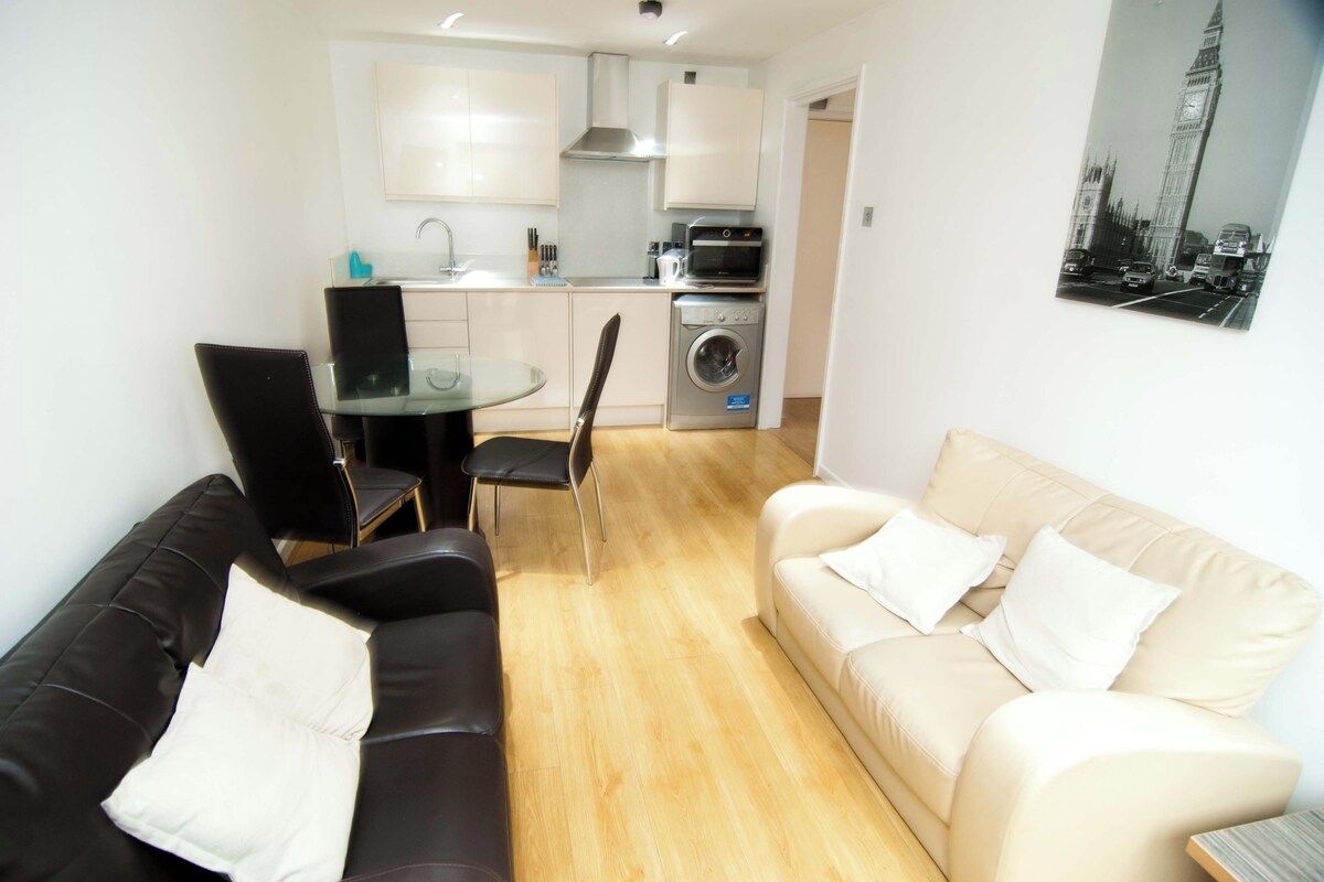 A modern living room with a black leather sofa, a beige sofa, and a glass dining table with chairs. A kitchen with white cabinets and appliances is visible in the background.