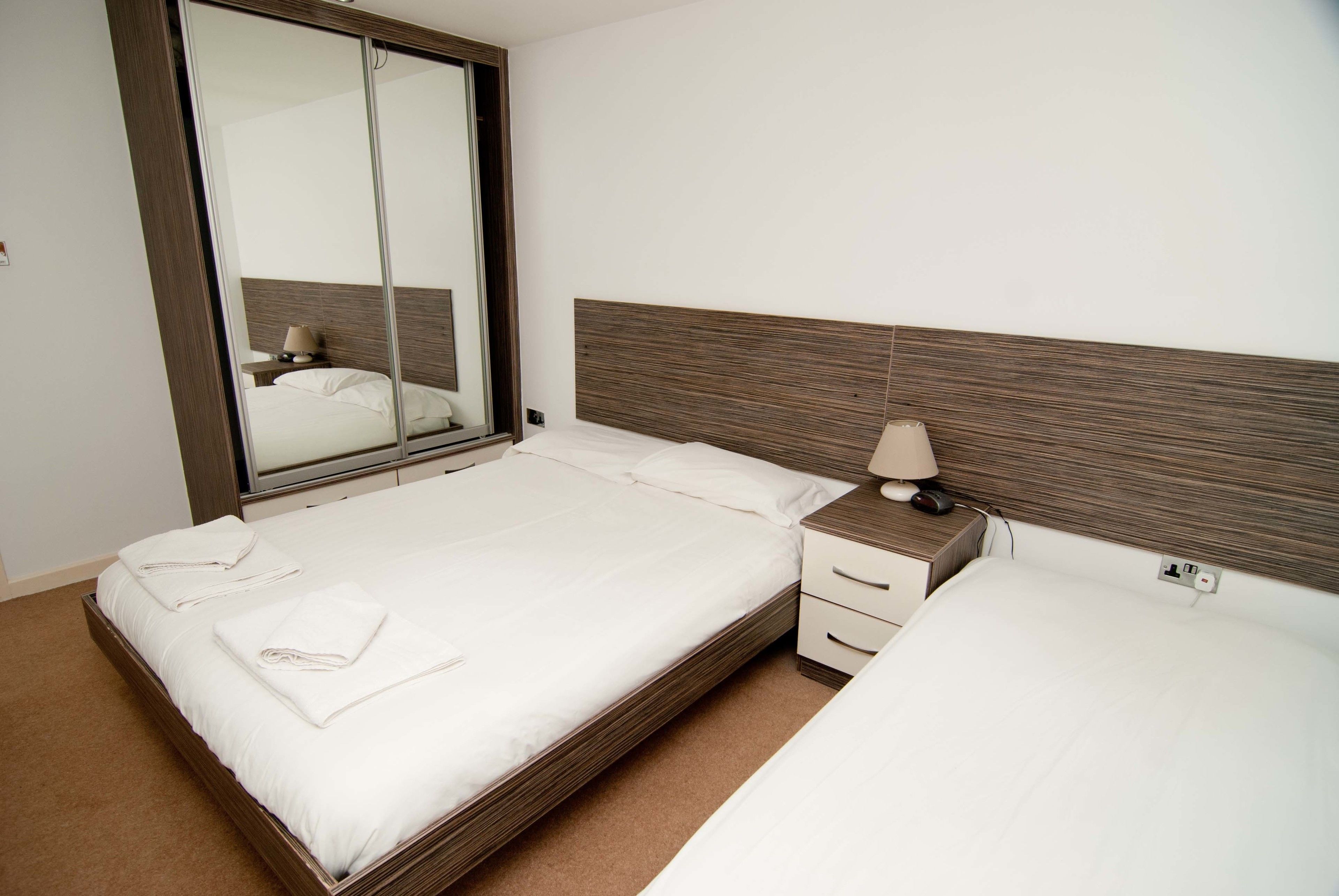 A hotel room with a large bed, a bedside table with a lamp, a mirror, and a wardrobe.