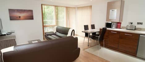 A modern living room with a sofa, a dining area with chairs, a kitchen with a microwave, and a view of the outdoors through large windows.