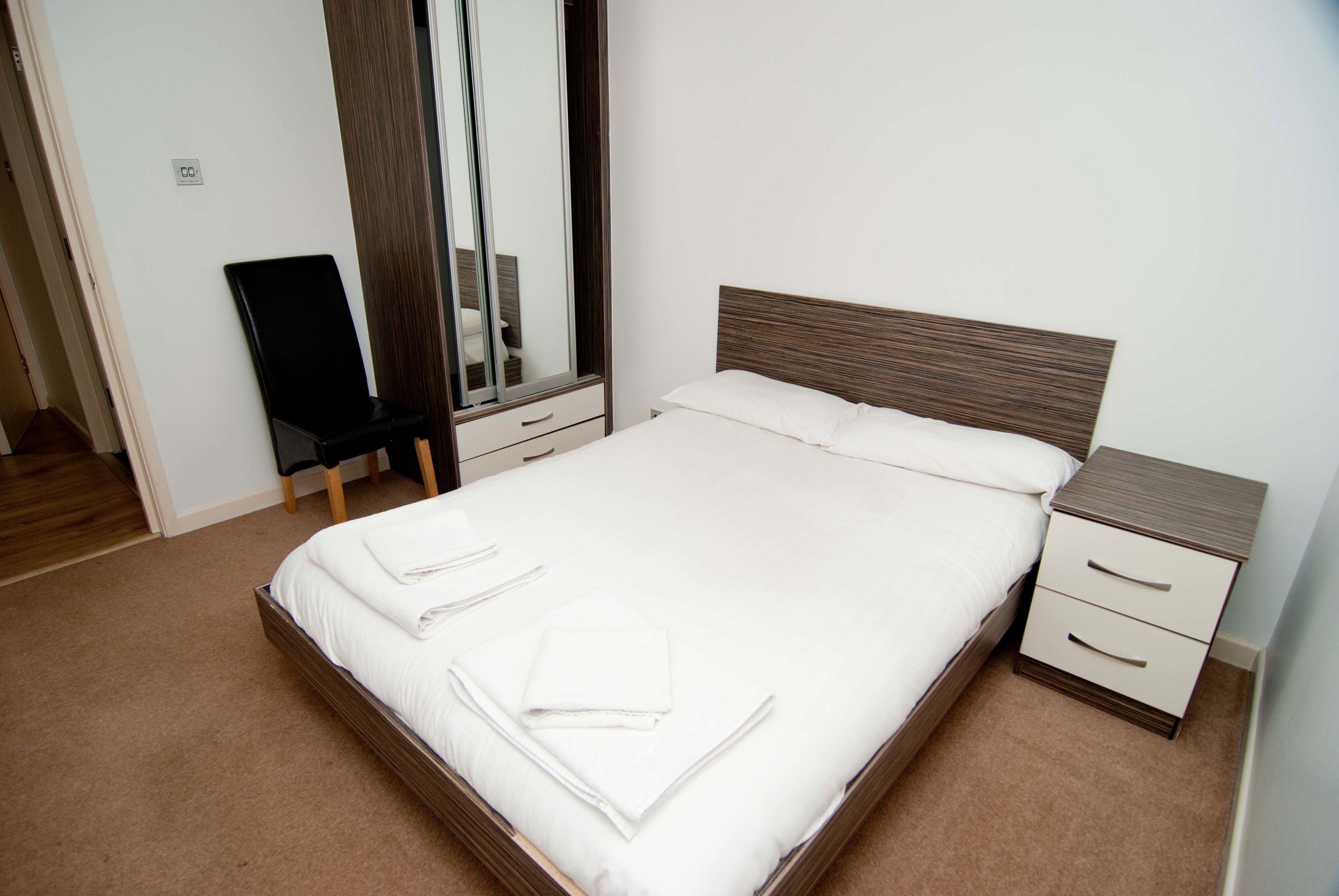 A neatly made bed with white linens and towels, a wooden headboard, a bedside table with drawers, and a mirror on the wall.