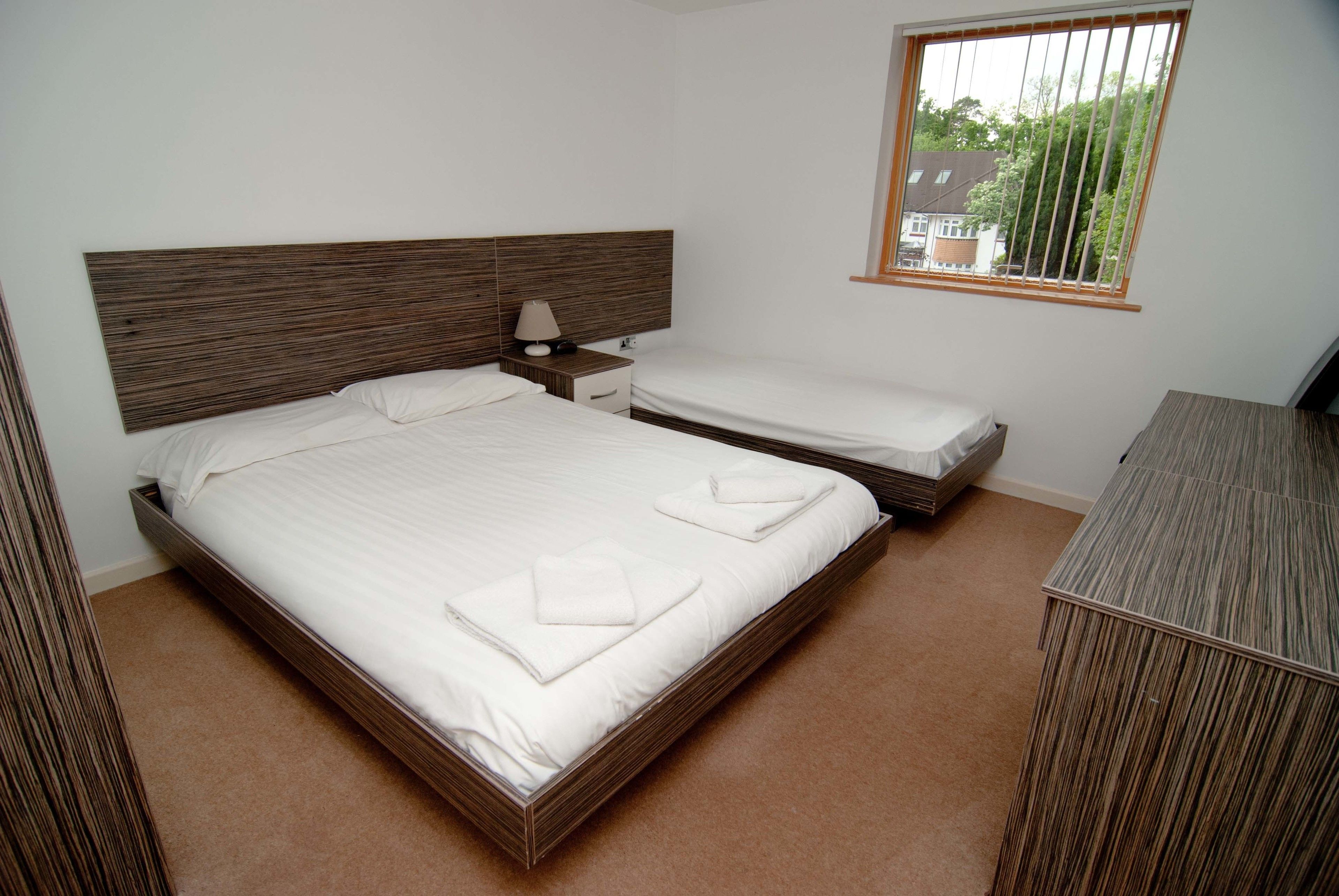 Een slaapkamer met een groot bed, een kleiner bed, een nachtkastje, een lamp, een dressoir en een raam met uitzicht op bomen en huizen.