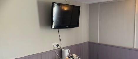 Una habitación con un televisor montado, una mesita con mantel blanco y un hervidor.