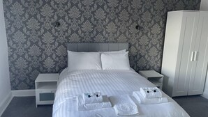A neatly made bed with white linens and towels, a grey patterned wallpaper, and a white wardrobe. - The Farmers Arms (Richmond)