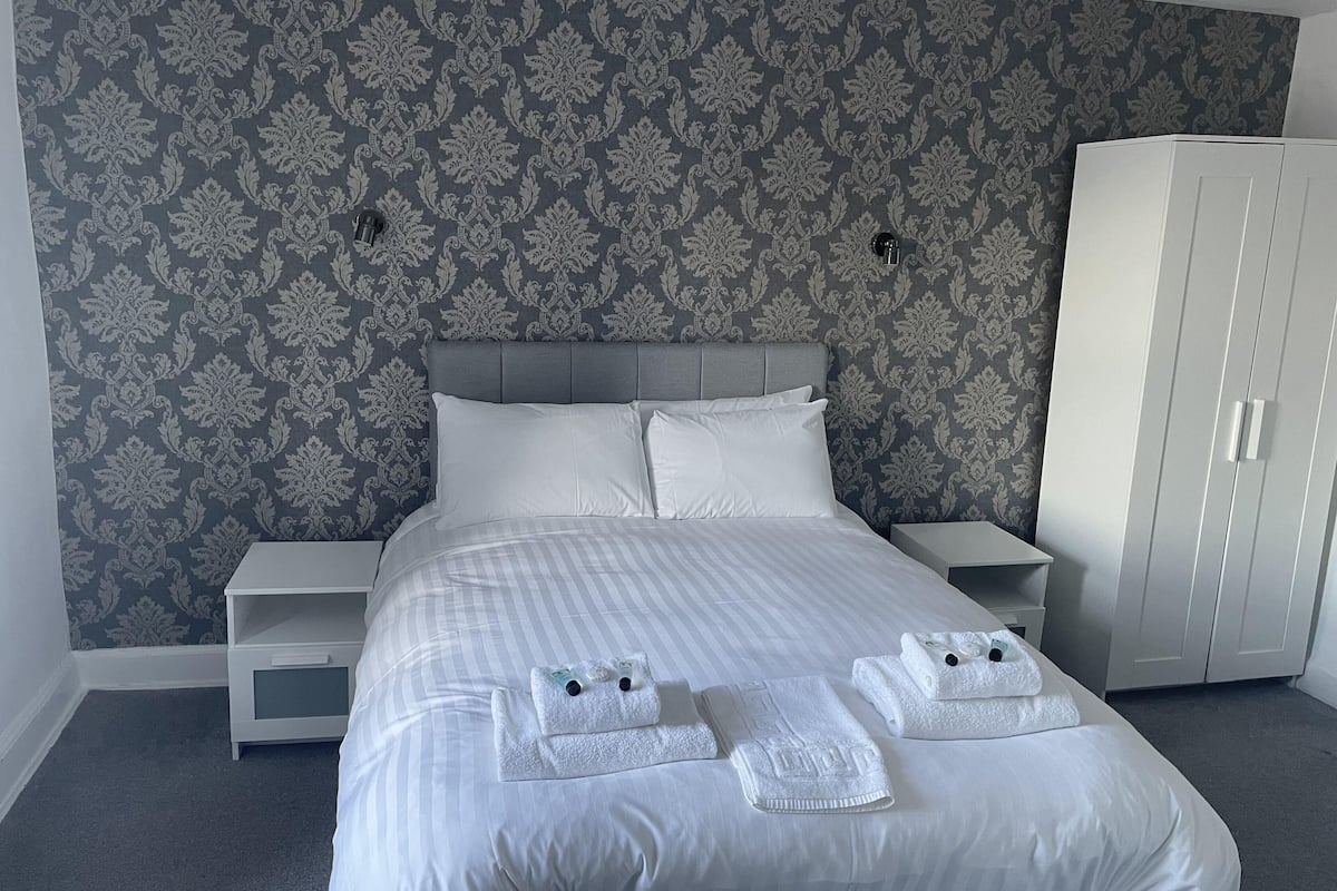 A neatly made bed with white linens and towels, a grey patterned wallpaper, and a white wardrobe.