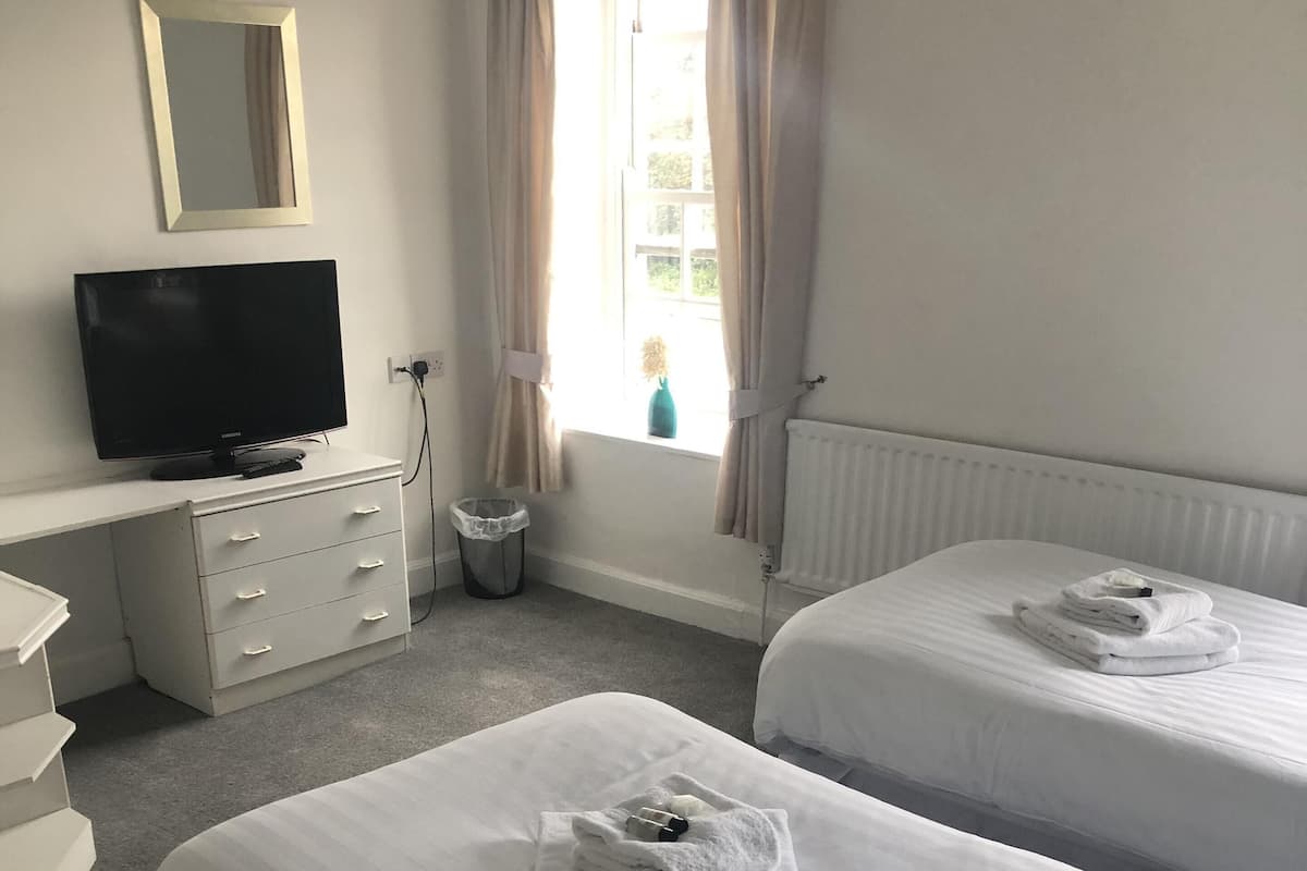 A hotel room with two beds, a TV, a dresser, a window with curtains, and a radiator.