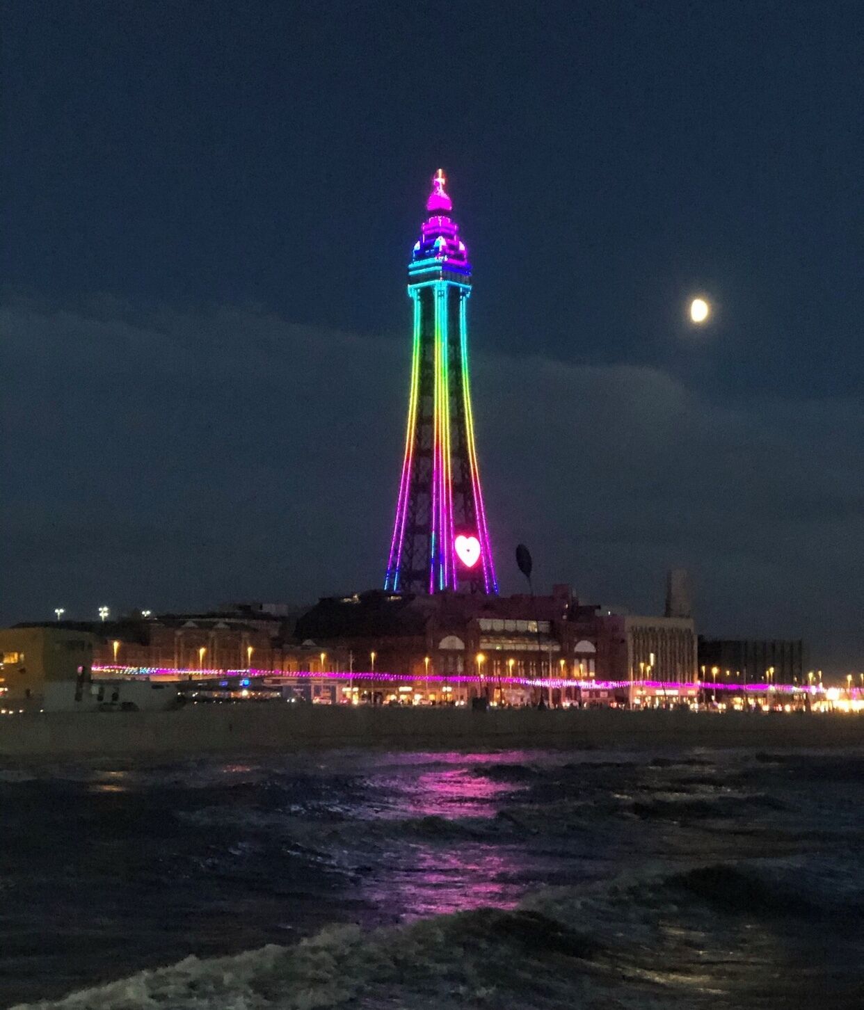 Uma torre alta com um pináculo, iluminada por luzes coloridas, e uma praia ao primeiro plano.